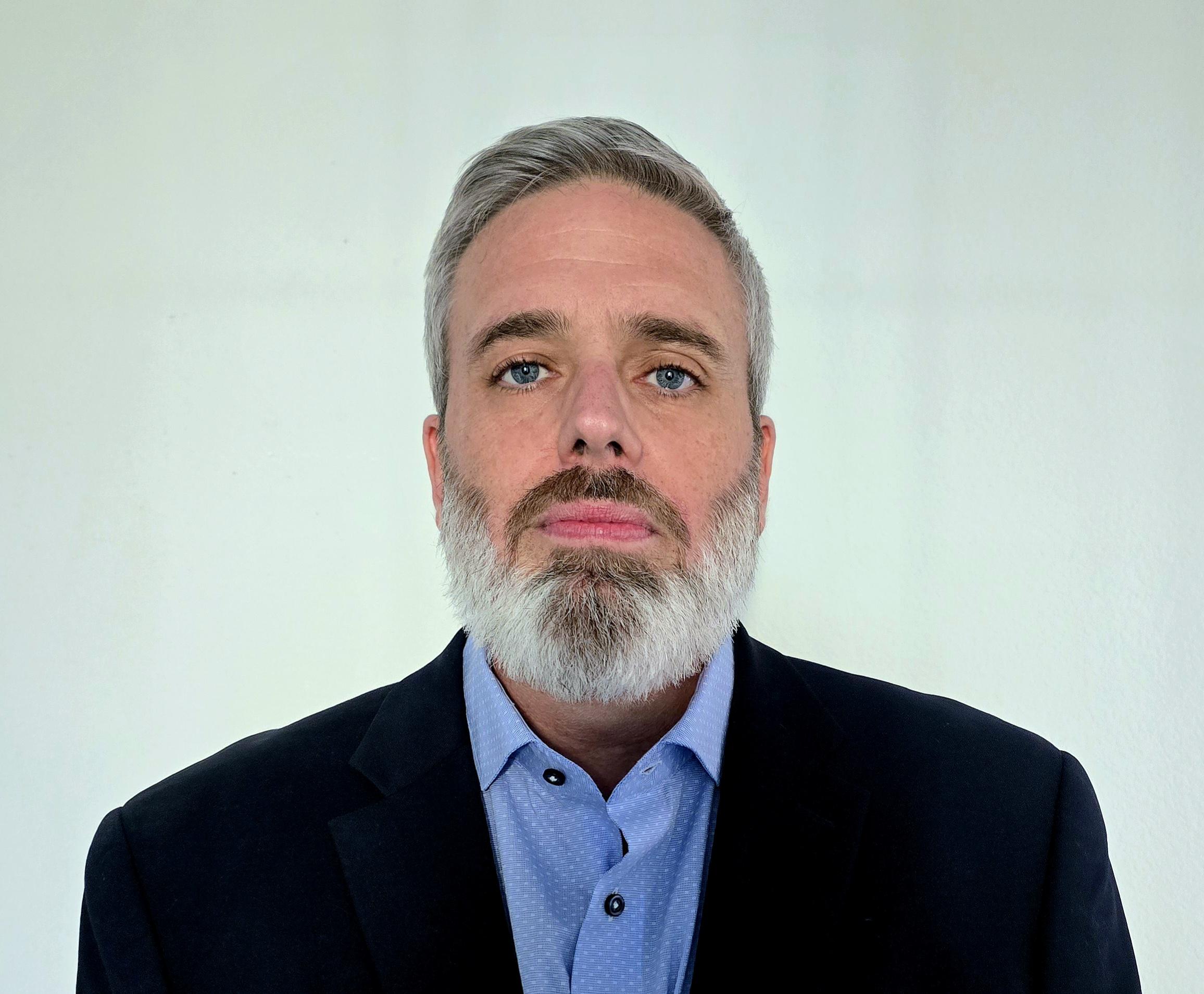 Recruiter Nate Shuman in a black suitcoat with a blue collared shirt. He has a short white beard, blue eyes, and a moustache.